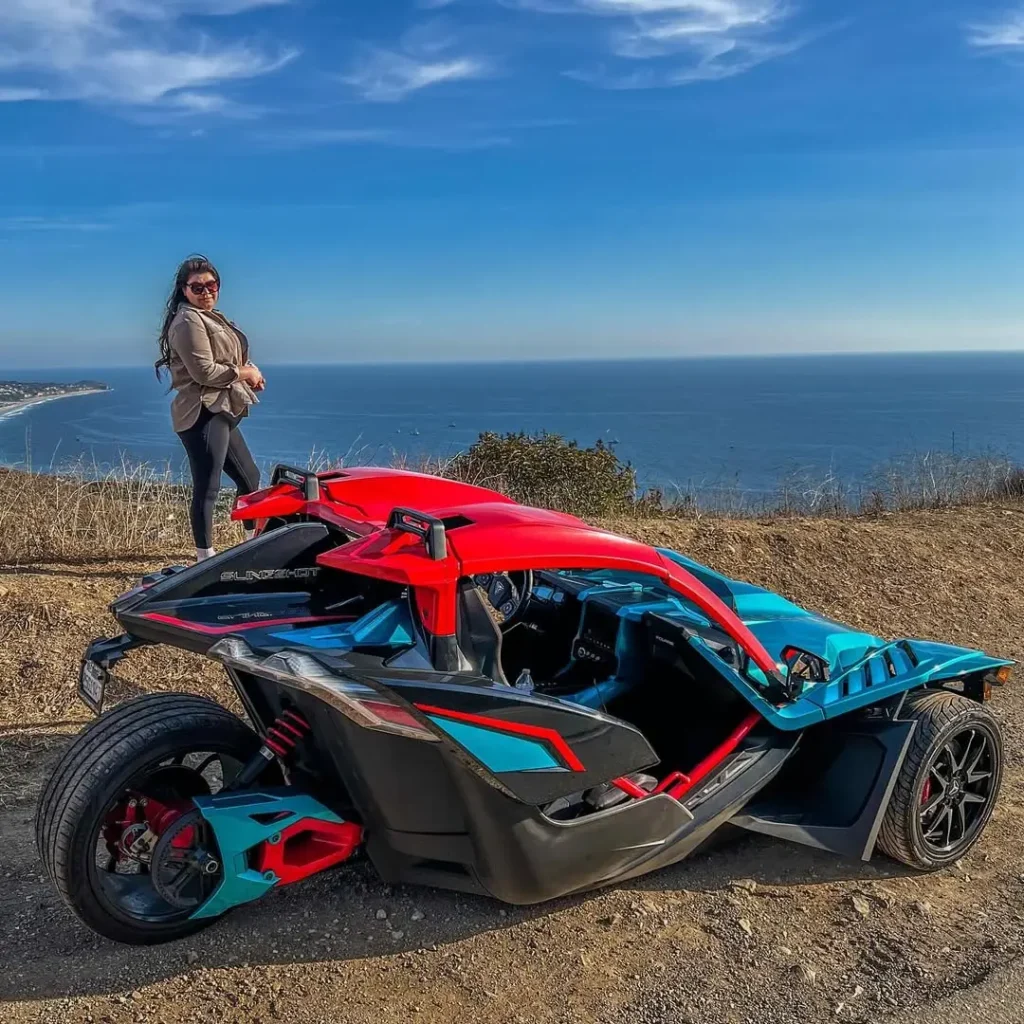 Person standing by a scenic coastal view behind a futuristic red and blue vehicle.